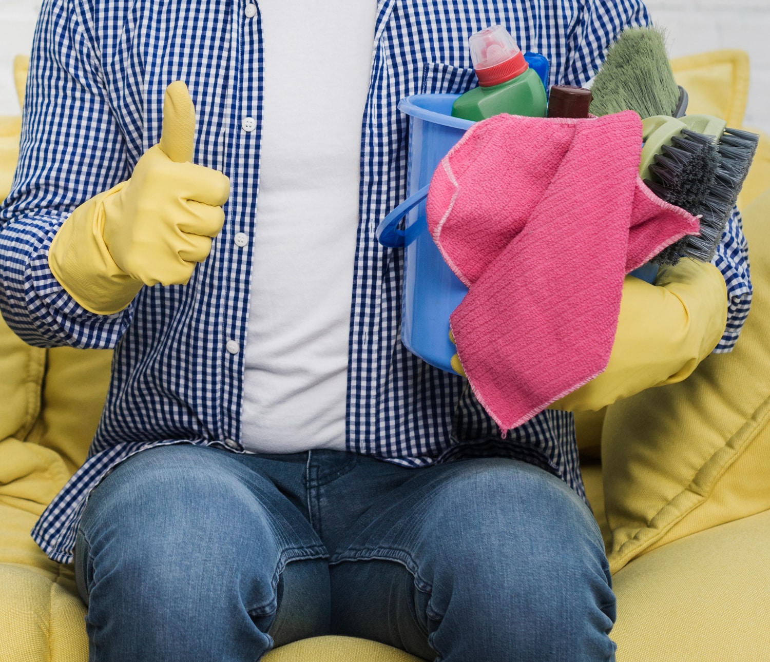 Homme assis sur un canapé jaune, tenant un seau rempli d'articles de nettoyage, faisant un pouce levé.