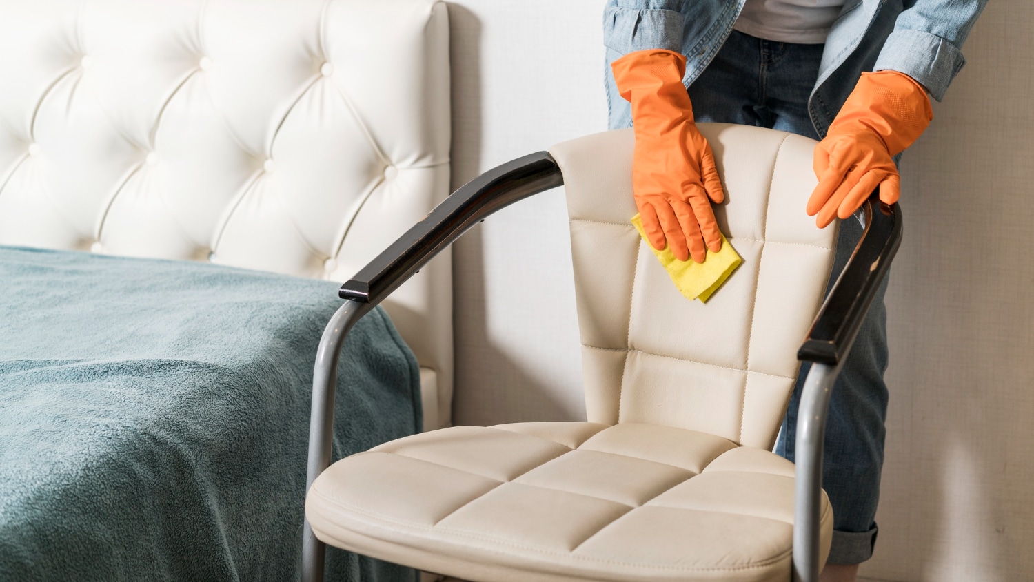 Personne en gants orange nettoyant avec soin un fauteuil en cuir crème à l'aide d'une éponge jaune.