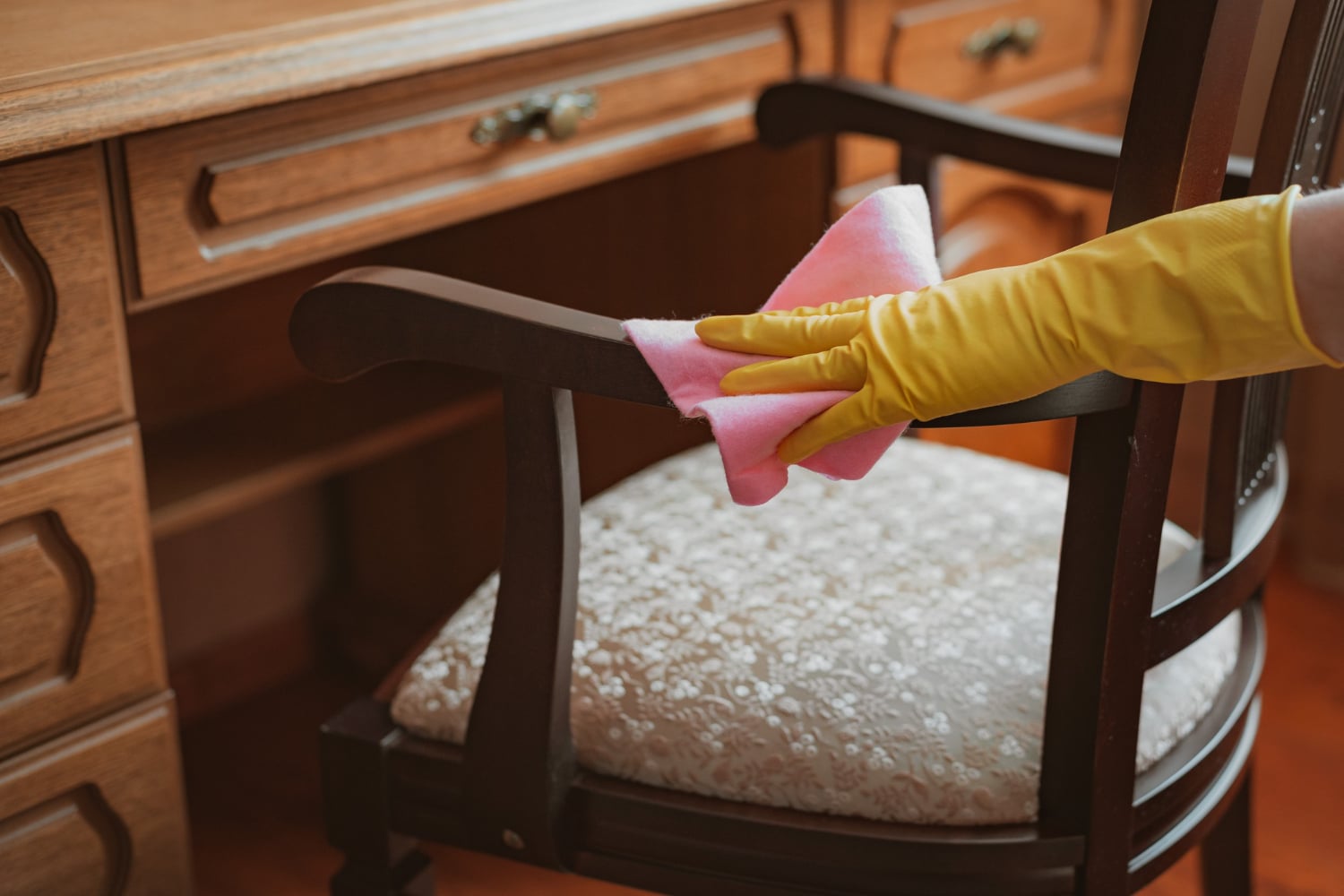 Main en gant jaune essuyant le bras en bois d'une chaise classique en tissu à motifs, à côté d'un bureau en bois.