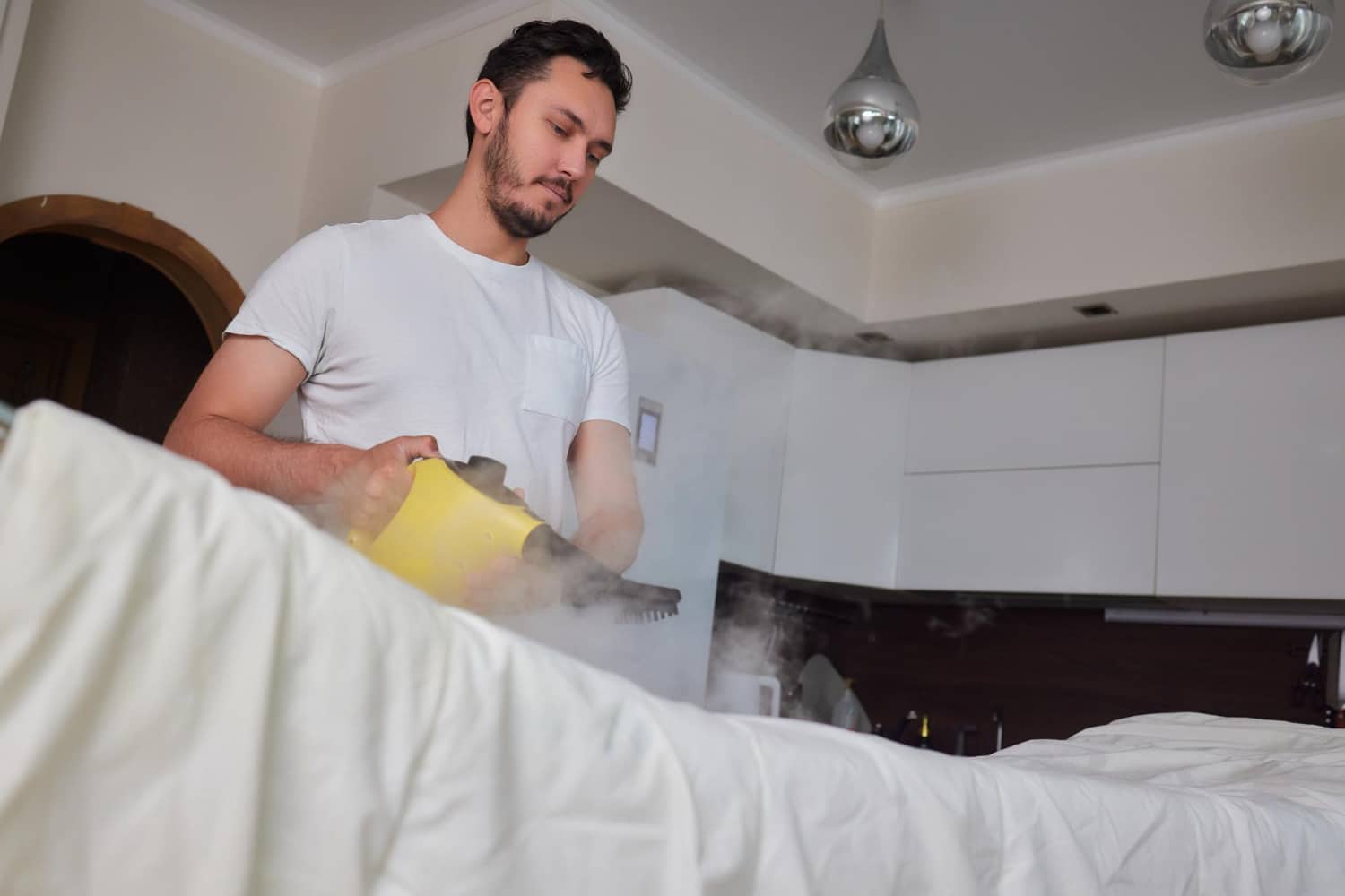 Homme utilisant un nettoyeur vapeur pour désinfecter un lit dans une chambre lumineuse.