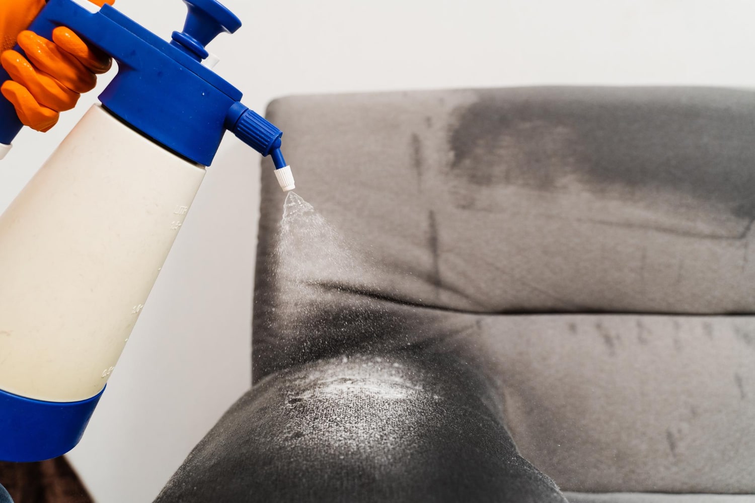 Gros plan sur une main gantée orange pulvérisant un produit de nettoyage sur un fauteuil gris foncé, faisant apparaître la mousse blanche du produit.