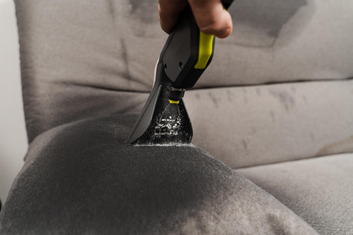 Gros plan sur une main utilisant un outil d'extraction pour nettoyer une tache sur un canapé en tissu gris.